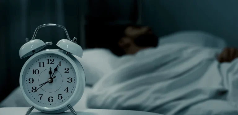 A white alarm clock showing 2:45 AM on a bedside table, with a man lying awake in bed, covered in a blanket, struggling with sleep in a dimly lit bedroom.